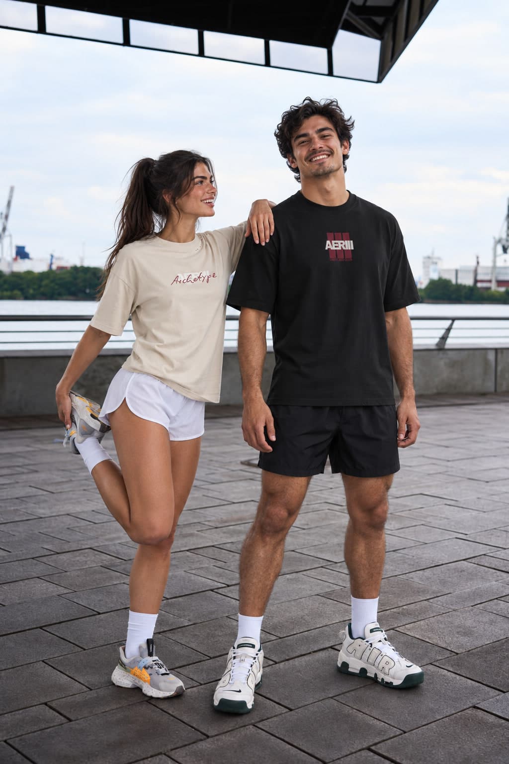 LIMITLESS CLUB BLACK tee in black on a male model, beside a woman in a pale AERIII tee, on a waterfront
